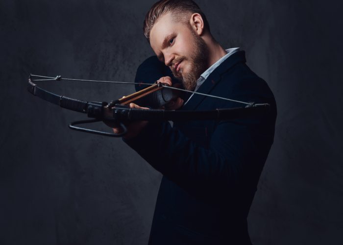 A serious, bearded male dressed in a suit holds crossbow.